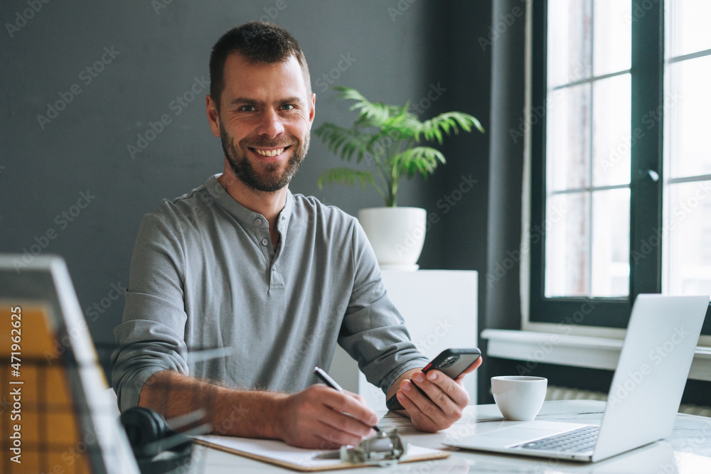 Young smiling business man in casual grey longsleeve working at laptop in dark modern office room Young smiling business man in casual grey longsleeve working at laptop in dark modern office room