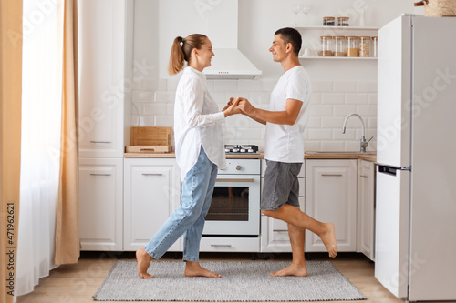 Side view portrait of beautiful couple dancing in kitchen at home, holding hands and looking at each other with love, expressing romantic feelings, celebrating anniversary.