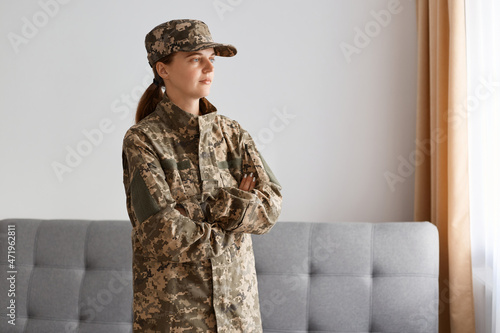Portrait of serious female airman standing indoor against gray sofa and window indoor, keeping hands folded and looking away, returning home after military serve.