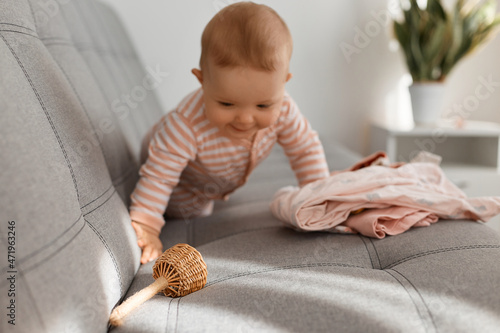 Cheerful baby girl plays with wooden toy and clothing at home. One-year-old child, crawling on the sofa with a toy in her hand, pose in the sunlight.