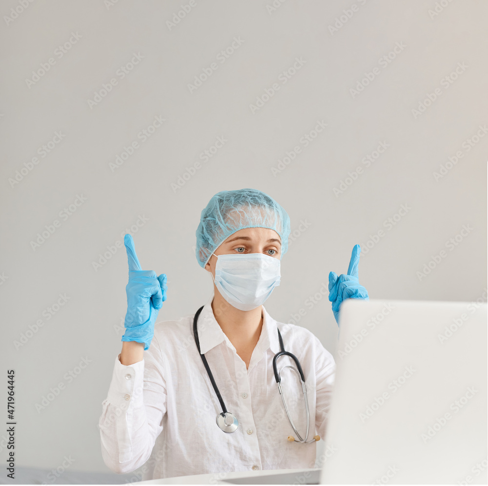 Indoor shot of attractive woman doctor sitting at table and working on laptop computer, has online video conference, pointing index fingers up, showing copy space. Indoor shot of attractive woman doctor sitting at table and working on laptop computer, has online video conference, pointing index fingers up, showing copy space.