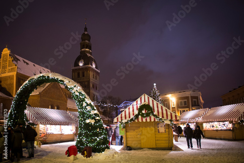Winter evening on the main square of the city, street lit by evening lights, before Christmas, New Year. Christmas Fair. Catholic cathedral, church, tower. Snowy streets. Riga, Latvia.