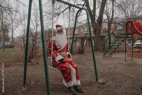 Fototapeta Naklejka Na Ścianę i Meble -  man dressed as Santa Claus with tangerines in a package sits on a children's swing against the backdrop of garages. The environmental problem of warming when there is no snow in winter.