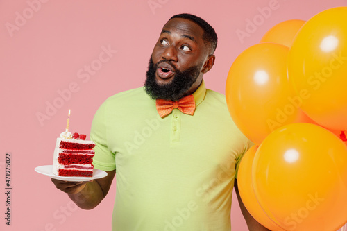Young happy black gay man in green t-shirt bow tie hold bunch of air inflated helium balloons celebrating birthday party sweet cake look aside on workspace isolated on plain pastel pink background © ViDi Studio
