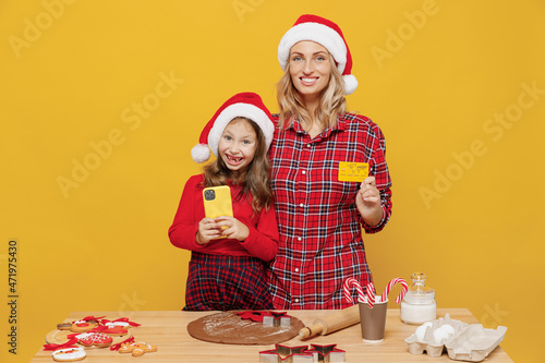 Two woman mother, fun child baby girl in red Christmas hat isolated on plain yellow background studio. Mom little kid cook ginger cookie at kitchen table home hold credit card. Happy New Year concept.