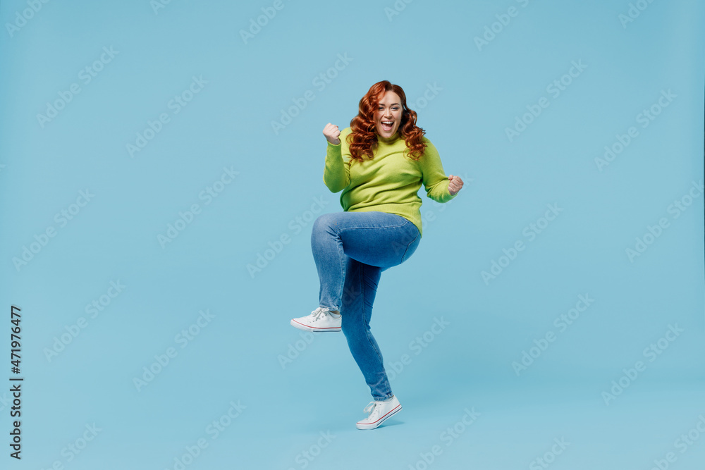 Full body overjoyed young chubby overweight plus size big fat fit woman wear green sweater do winner gesture raise up leg isolated on plain blue background studio portrait. People lifestyle concept. Full body overjoyed young chubby overweight plus size big fat fit woman wear green sweater do winner gesture raise up leg isolated on plain blue background studio portrait. People lifestyle concept.