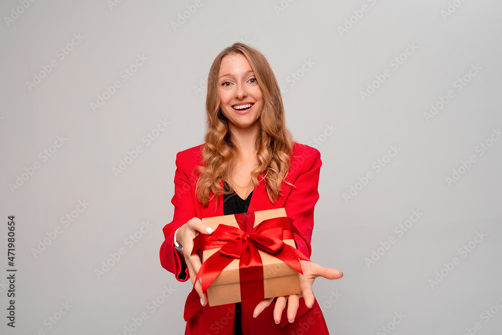 happy smiling young woman holding gift box decorated with red bow. Attractive blonde girl in stylish red jacket posing isolated on gray background. New Year Women's Day birthday holiday concept happy smiling young woman holding gift box decorated with red bow. Attractive blonde girl in stylish red jacket posing isolated on gray background. New Year Women's Day birthday holiday concept