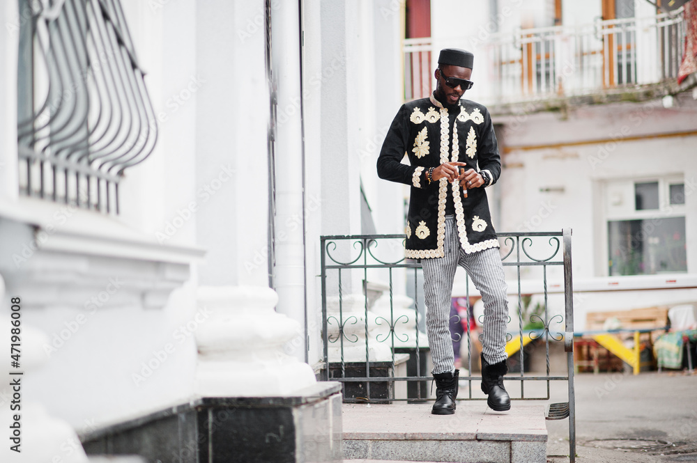 Mega stylish african man in traditional jacket pose. Fashionable black guy in hat and sunglasses with cigar in hand.
