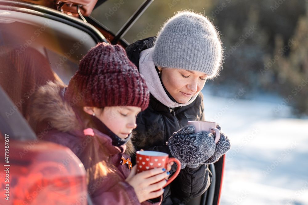 Family vacation, family values. Mother spends fun with her daughter in the winter in the park. Mom and daughter are drinking tea while sitting in the open trunk of their SUV.