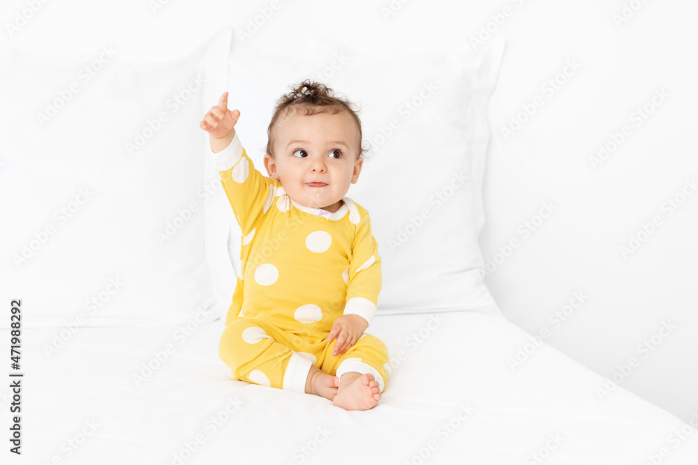 Cute baby sitting on white bed raising finger in the air Cute baby sitting on white bed raising finger in the air