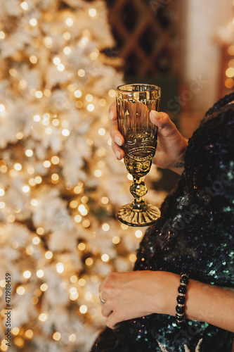woman in shiny dress holds green drink glass in her hand against background of Christmas tree and bokeh. selective focus