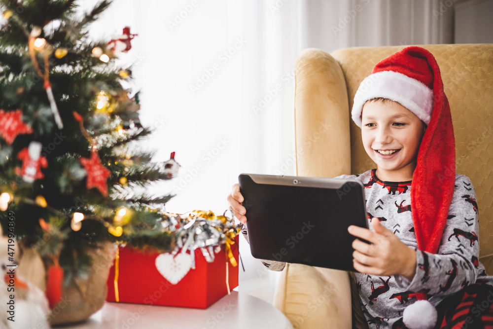 Happy teenager boy in pajamas and red santa hat with tablet having video chat at home. Smiling child sitting on armchair watching video at laptop. Christmas tree with decorative toys and lights.