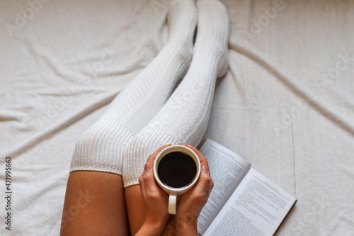 Female hands holding a coffee cup on legs in woolen winter socks in a cozy bed with a book at home. Close up, selective focus