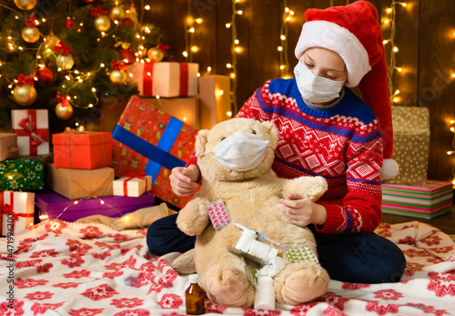 Child girl as Santa helper posing in new year decoration. Wearing a protective face mask against viruses, coronaviruses. She plays with a toy bear, treats him, measures his temperature