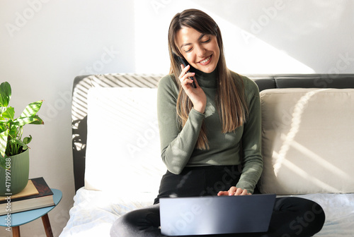 Young woman sits on a bed, writes on a laptop and talks on the phone.
Beautiful woman sitting on a bed occupied by a laptop and a telephone.