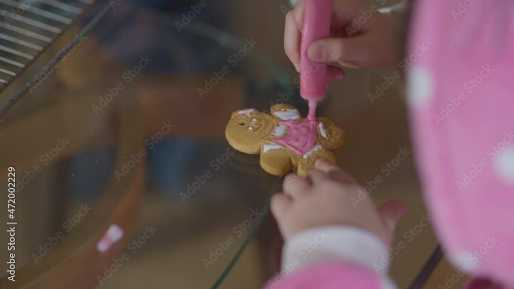 Close-up gingerbread cookie with little female hands decorating dessert with pink royal icing. Unrecognizable Caucasian little girl preparing sweet tasty traditional Christmas food indoors Close-up gingerbread cookie with little female hands decorating dessert with pink royal icing. Unrecognizable Caucasian little girl preparing sweet tasty traditional Christmas food indoors