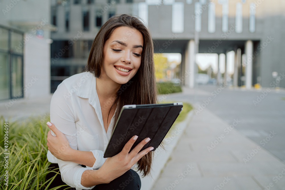 Break from office, smartly dressed girl sits on concrete outside in front of glass modern corporate building, ornamental plants around her, woman is holding a tablet, watching videos, series Break from office, smartly dressed girl sits on concrete outside in front of glass modern corporate building, ornamental plants around her, woman is holding a tablet, watching videos, series