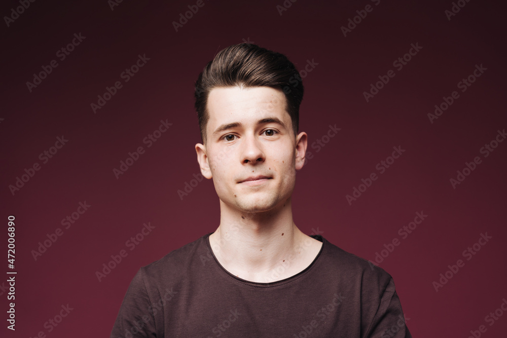 Fototapeta premium Isolated shot of young serious handsome male with trendy hairdo, wears casual brown t shirt, has serious expression as listens to interlocutor, poses in studio against crimson background