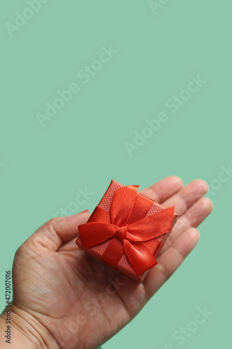 Mão de uma pessoa segurando um presente pequeno, embrulhado com papel e laço vermelhos com fundo verde claro. photo