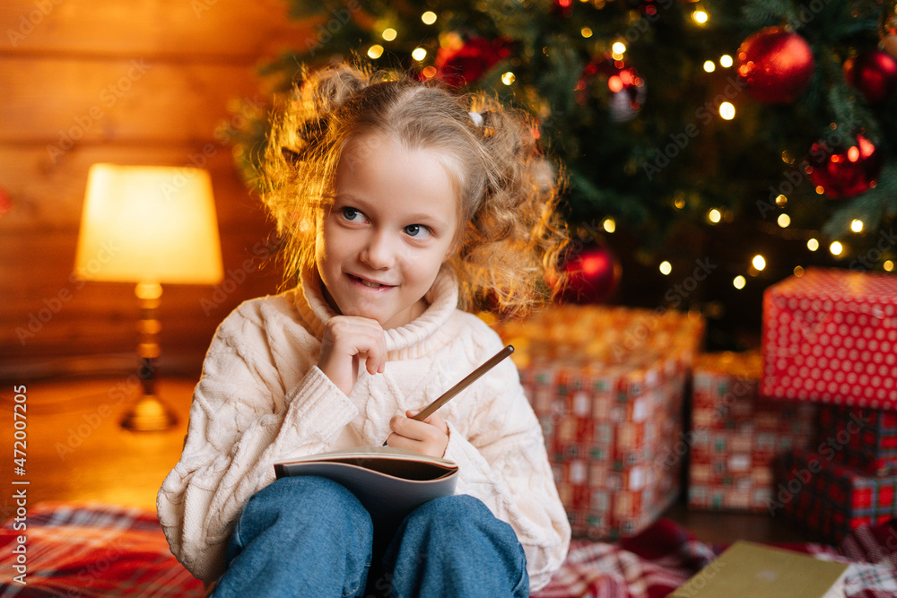 Close-up portrait of pensive cheerful little blonde girl writing letter to Santa Claus sitting on floor on background of Christmas tree and gift boxes, in living room with festive bright interior. Close-up portrait of pensive cheerful little blonde girl writing letter to Santa Claus sitting on floor on background of Christmas tree and gift boxes, in living room with festive bright interior.