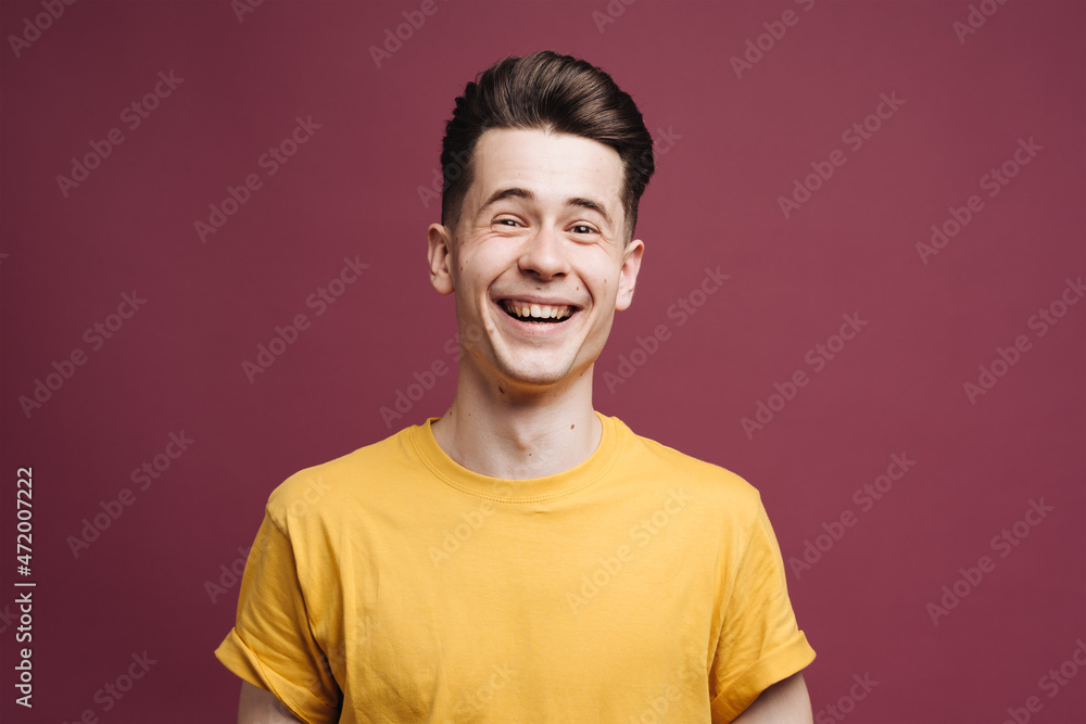 Isolated shot of young laughing handsome male with trendy hairdo, wears casual brown t shirt, has serious expression as listens to interlocutor, poses in studio against crimson background Isolated shot of young laughing handsome male with trendy hairdo, wears casual brown t shirt, has serious expression as listens to interlocutor, poses in studio against crimson background