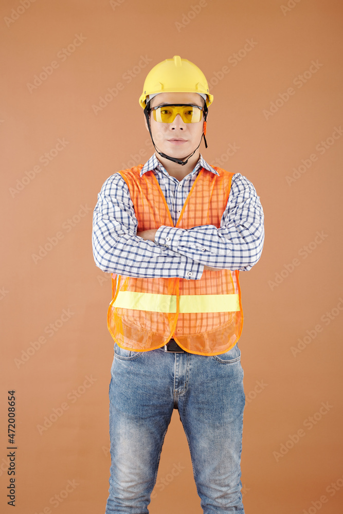 Portrait of confident contractor in bright vest, goggles and hardhat crossing arms and looking at camera