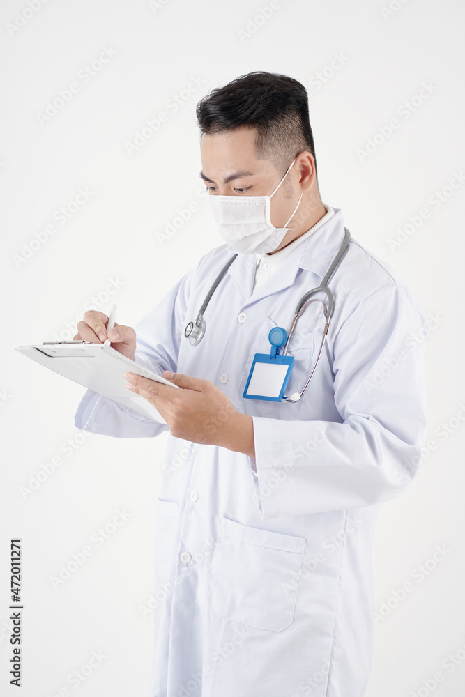 Male general practitioner in medical mask filling document on clipboard Male general practitioner in medical mask filling document on clipboard