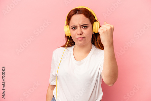 Young caucasian woman listening to music isolated on pink background showing fist to camera, aggressive facial expression.