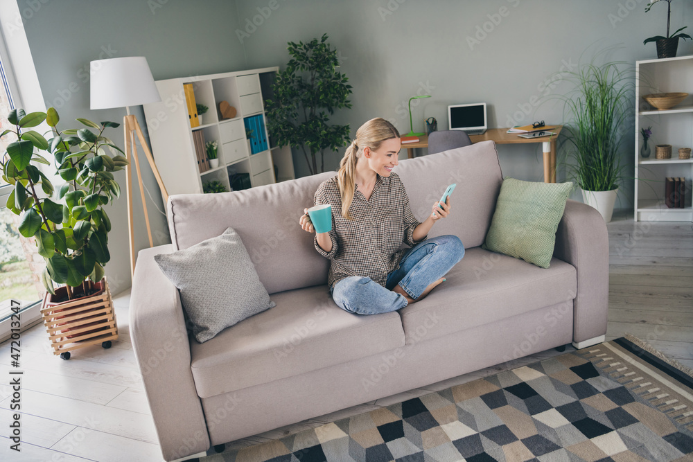 Full length photo of positive modern lady sit divan use app smart phone hold cacao cup read quarantine news in apartment room Full length photo of positive modern lady sit divan use app smart phone hold cacao cup read quarantine news in apartment room
