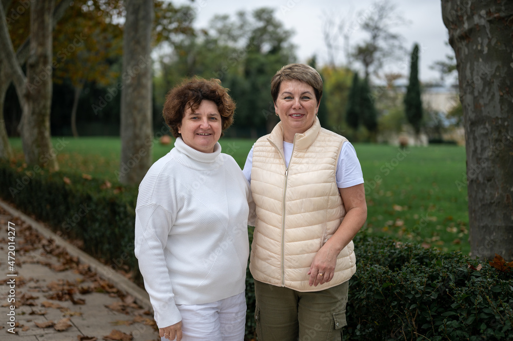 Two older women standing outside and looking at camera. two middle-aged women walking in park Two older women standing outside and looking at camera. two middle-aged women walking in park