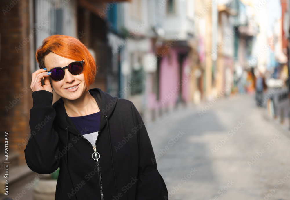 Young lesbian woman confidently walks down street and looks ahead through sunglasses. Young cheerful woman with bright red hair walks down narrow street Young lesbian woman confidently walks down street and looks ahead through sunglasses. Young cheerful woman with bright red hair walks down narrow street