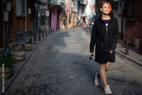Young lesbian woman confidently walks down the street and looks ahead. Young cheerful woman with bright red hair walks down a narrow street