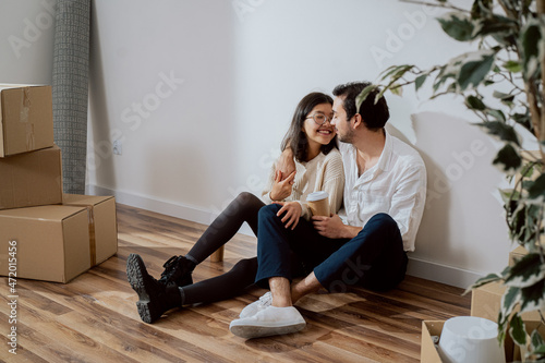 A couple in love are sitting by a wall in a new apartment, a gallant man dressed in a white shirt embraces a smiling pretty woman, they are drinking coffee, flirting, around unpacked boxes of things