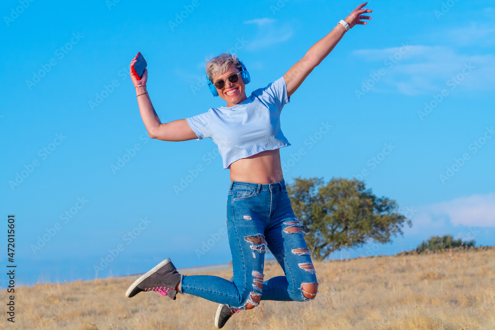 Smiling pretty young woman with short blonde hair, blue helmets, belly button t-shirt and ripped jeans jumping and laughing with her smart phone in hand. lifestyle concept Smiling pretty young woman with short blonde hair, blue helmets, belly button t-shirt and ripped jeans jumping and laughing with her smart phone in hand. lifestyle concept
