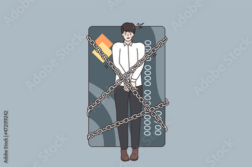 Financial crisis and debts concept. Young stressed man being locked with chains to huge credit card having debts as slave over grey background 