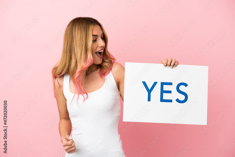 Young woman over isolated pink background holding a placard with text YES and celebration a victory Young woman over isolated pink background holding a placard with text YES and celebration a victory