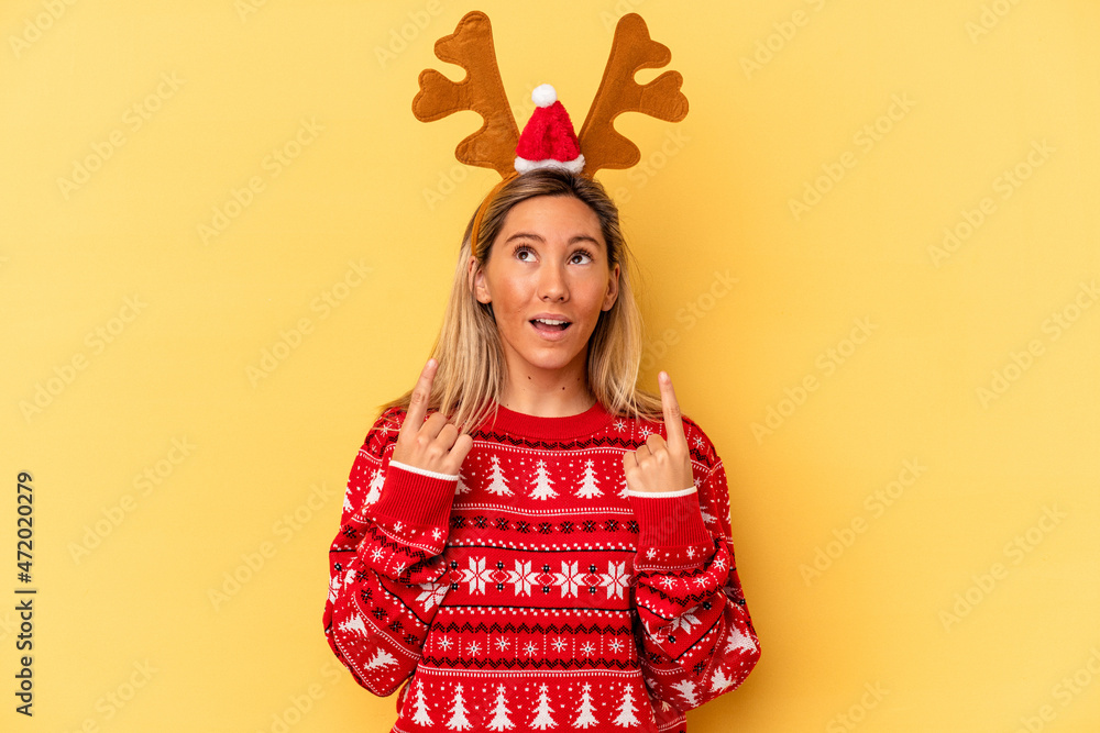 Young caucasian woman wearing a christmas reindeer hat isolated on beige background pointing upside with opened mouth. Young caucasian woman wearing a christmas reindeer hat isolated on beige background pointing upside with opened mouth.