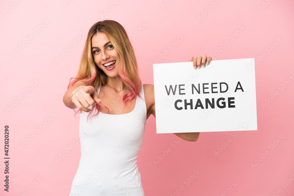 Young woman over isolated pink background holding a placard with text We Need a Change and pointing to the front Young woman over isolated pink background holding a placard with text We Need a Change and pointing to the front