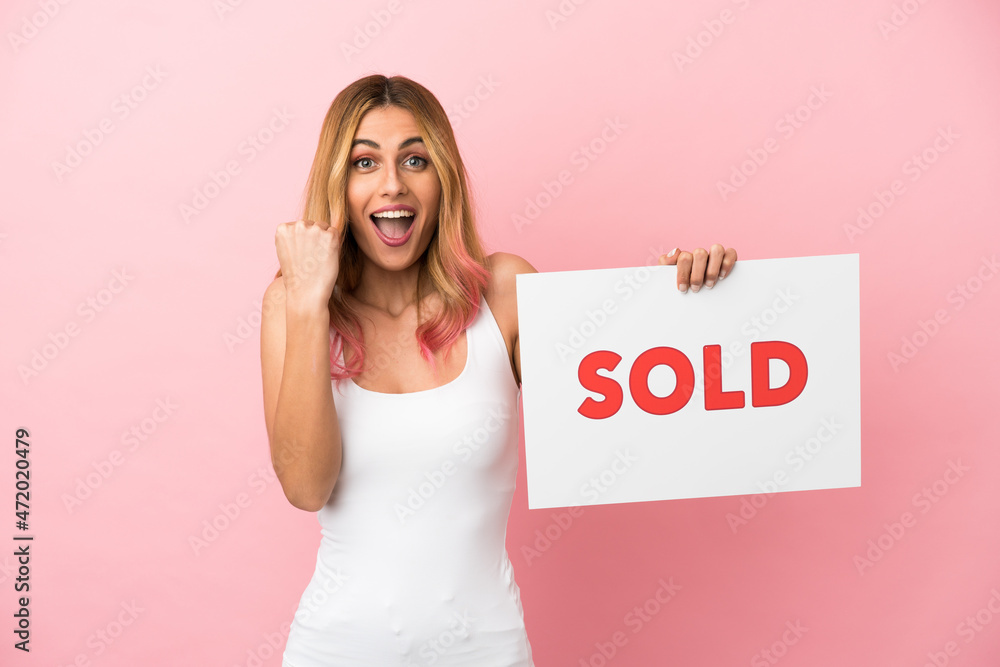 Young woman over isolated pink background holding a placard with text SOLD and celebrating a victory Young woman over isolated pink background holding a placard with text SOLD and celebrating a victory