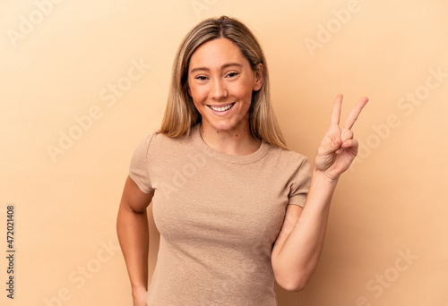 Young caucasian woman isolated on beige background showing victory sign and smiling broadly.