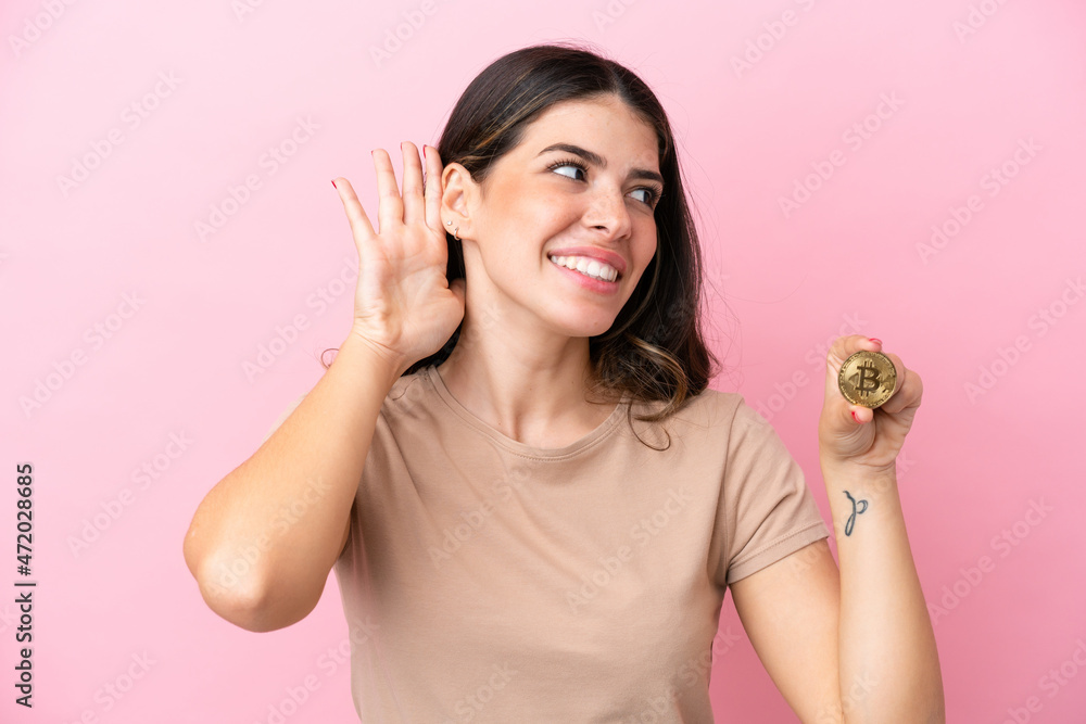 Young Italian woman holding a Bitcoin isolated on pink background listening to something by putting hand on the ear Young Italian woman holding a Bitcoin isolated on pink background listening to something by putting hand on the ear