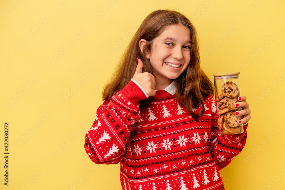 Little caucasian girl holding a Christmas cookies isolated on yellow background smiling and raising thumb up Little caucasian girl holding a Christmas cookies isolated on yellow background smiling and raising thumb up
