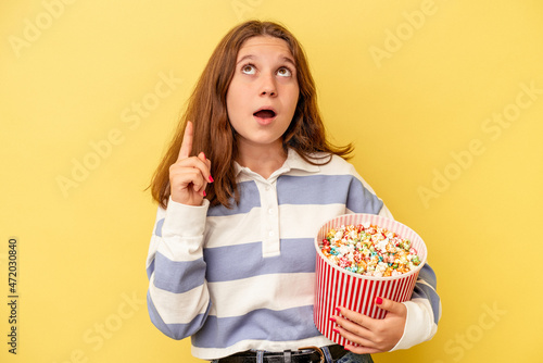 Little caucasian girl holding popcorns isolated on yellow background pointing upside with opened mouth.