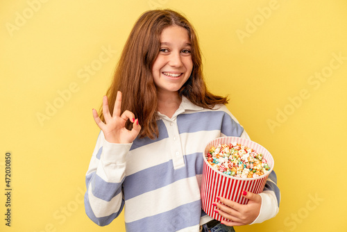 Little caucasian girl holding popcorns isolated on yellow background cheerful and confident showing ok gesture.