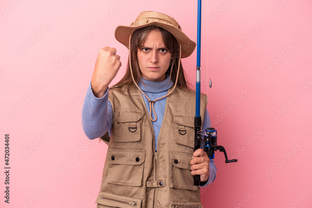 Young caucasian fisherwoman holding a rod isolated on pink background showing fist to camera, aggressive facial expression. Young caucasian fisherwoman holding a rod isolated on pink background showing fist to camera, aggressive facial expression.