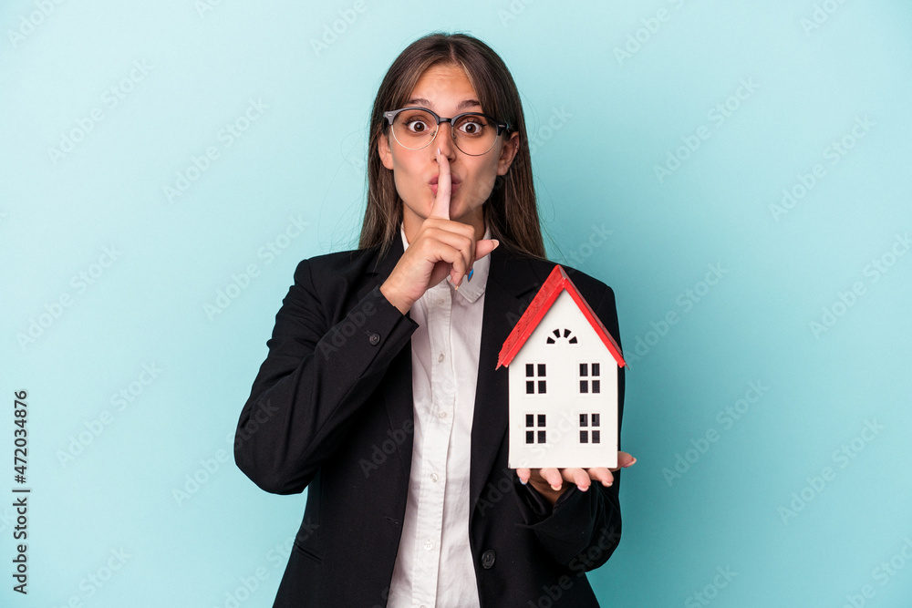 Young business woman holding a toy home isolated on blue background keeping a secret or asking for silence. Young business woman holding a toy home isolated on blue background keeping a secret or asking for silence.