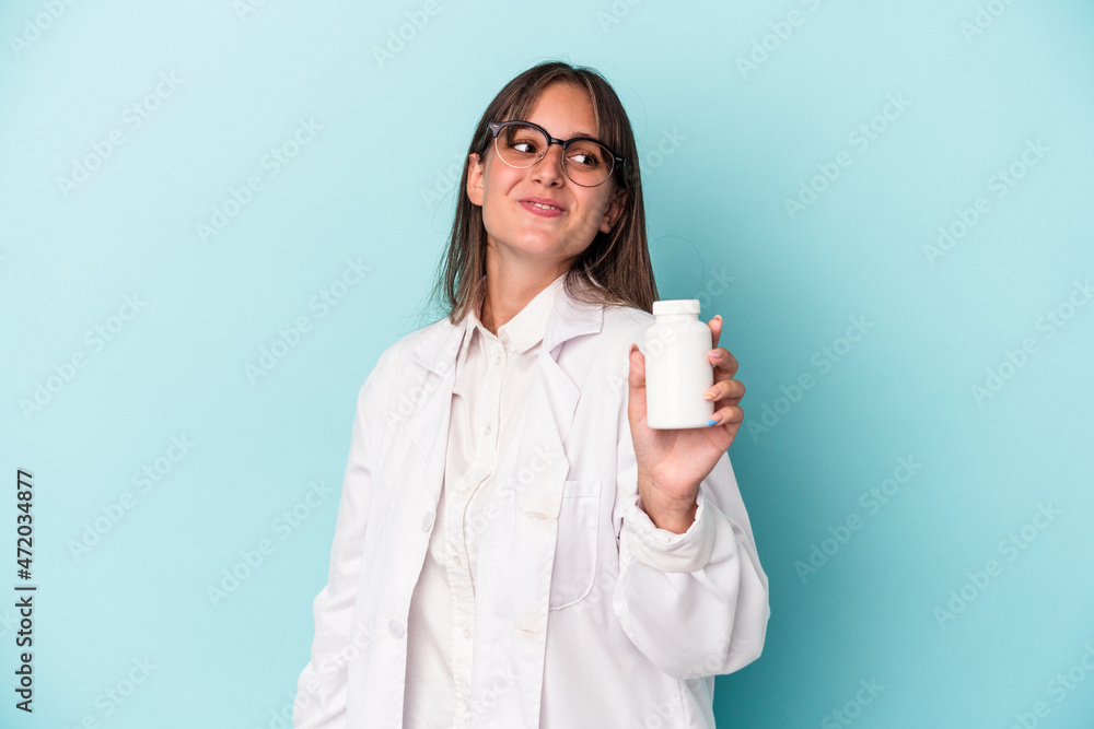 Young pharmacist woman holding pills isolated on blue background looks aside smiling, cheerful and pleasant. Young pharmacist woman holding pills isolated on blue background looks aside smiling, cheerful and pleasant.