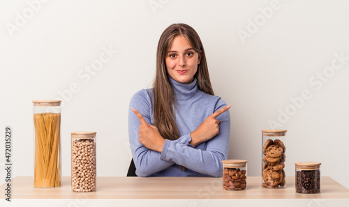 Young caucasian woman sitting at a table with food pot isolated on white background points sideways, is trying to choose between two options.