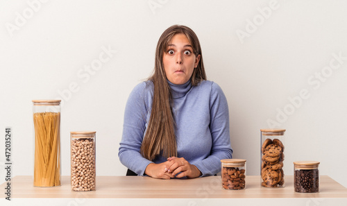 Young caucasian woman sitting at a table with food pot isolated on white background shrugs shoulders and open eyes confused.