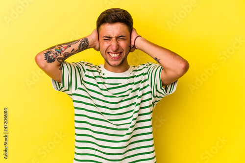 Young caucasian man with tattoos isolated on yellow background  covering ears with hands.